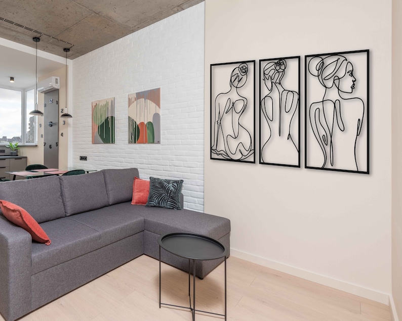 May include: Three black metal wall sculptures depicting abstract line drawings of women. The sculptures are mounted on a white brick wall in a modern living room.