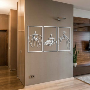 May include: Three white metal wall art pieces with line drawings of people holding cups of coffee or tea. The art is framed in a thin white frame and hung on a beige wall.