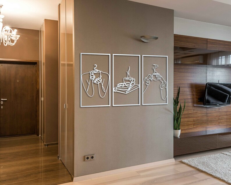 May include: Three white metal wall art pieces with line drawings of people holding cups of coffee or tea. The art is framed in a thin white frame and hung on a beige wall.