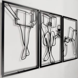 May include: Three-panel black metal wall art depicting figures reading and relaxing. One panel shows legs and a book, another a person with a glass of wine, and the third a person with a book. The art is mounted on a white wall.
