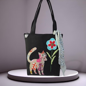 May include: A black tote bag featuring a beige cat appliqué with colorful patterned patches and a large blue and red flower appliqué. The bag has two black shoulder straps, a silver zipper pull, and a light blue floral patterned fabric ribbon tied.