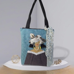 Op de afbeelding: Een blauwe tote bag met zwarte schouderbanden, voorzien van een applicatie van een persoon met een hoed met brede rand, een parelketting en een donkerbruine top met stippen en witte kanten rand. Een bloemrijke stoffen strook hangt aan de zijkant.