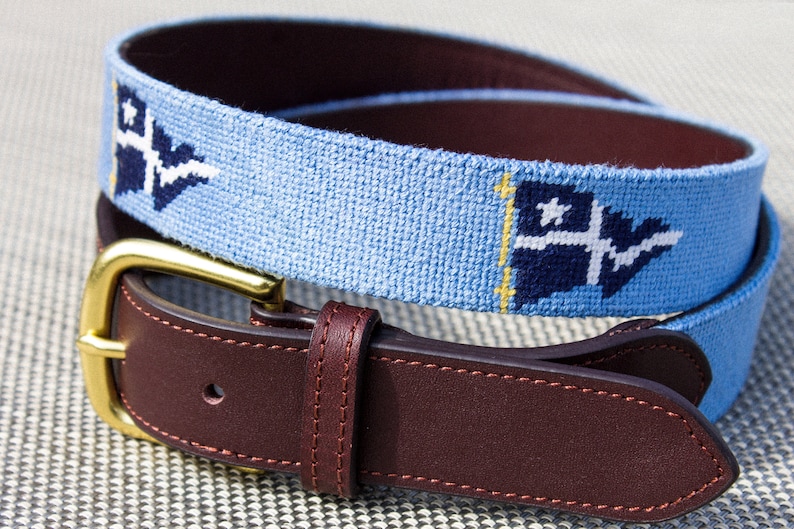 Custom needlepoint belt by Fish Creek brands. image displays a baby blue belt with navy blue burgees. Additionally the image shows a cowhide leather backing and solid brass buckle.
