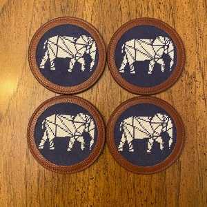 May include: Set of four round coasters with a brown leather border and a blue fabric center. Each coaster features a white embroidered elephant design.
