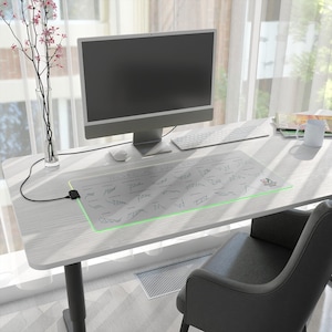 Op de afbeelding: Een witte bureau met een computer, toetsenbord en muis. Een grote, rechthoekige muismat met een groen verlichte rand en abstracte ontwerpen ligt op het bureau. Een vaas met roze bloemen staat links.