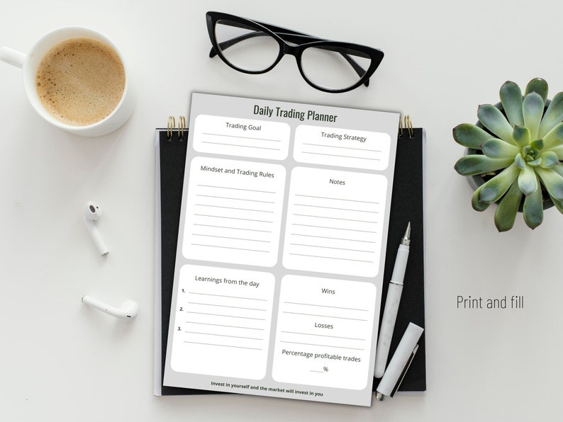 May include: A daily trading planner with sections for goals, strategy, notes, and daily learnings. The planner is on a black spiral notebook, with a pen, glasses, a cup of coffee, and a succulent plant. The text "Print and fill" is visible.