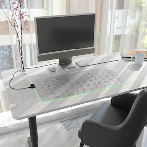 Op de afbeelding: Een witte bureau met een computer, toetsenbord en muis. Een grote, rechthoekige muismat met een groen verlichte rand toont een financieel diagram. Een vaas met roze bloemen staat links van de computer.