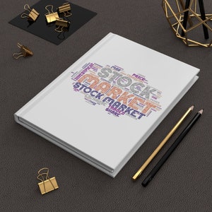 May include: A white notebook featuring a word cloud design with the words "STOCK MARKET" on the cover. The notebook is on a dark surface with gold and black pencils, and gold binder clips.