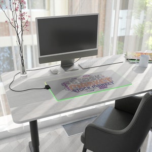 May include: A rectangular mouse pad with the words "STOCK MARKET" in a word cloud design. The pad has a green illuminated border and is on a white desk with a computer, keyboard, and mouse. A black chair is in the foreground.