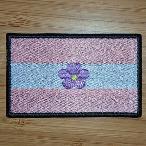 May include: A pink, blue, and white embroidered patch with a purple flower with a yellow center. The patch has a black border.