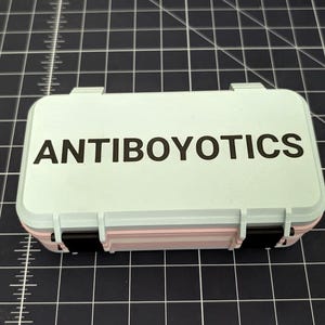May include: A small, rectangular, light blue case with the word "ANTIBOYOTICS" printed in black on the lid. The case has a pink and black accent and is closed with two black clasps. The case is on a black grid surface.