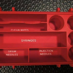 May include: A red organiser with compartments labelled for medical supplies. The organiser has sections for clean wipes, syringes, draw needles, and injection needles. It also has several circular compartments.