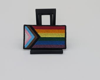 May include: A rectangular embroidered patch featuring the Progress Pride flag design. The patch displays horizontal stripes in red, orange, yellow, green, blue, and purple, with a chevron on the left side containing light blue, pink, white, brown, and black.