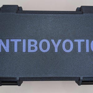 May include: A dark gray rectangular case with the word "ANTIBOYOTICS" in blue lettering. The case has a textured surface and black latches on the front. The case is closed.