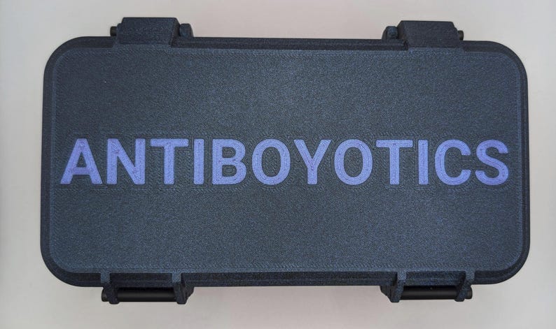 May include: A dark gray rectangular case with the word "ANTIBOYOTICS" in blue lettering. The case has a textured surface and black latches on the front. The case is closed.