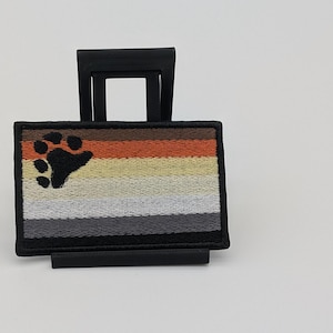May include: A rectangular embroidered patch featuring horizontal stripes in shades of orange, tan, white, gray, and black. A black paw print is stitched onto the left side of the patch. The patch is displayed on a small black plastic stand.