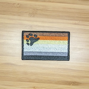May include: A brown, orange, beige, gray, and black embroidered patch with a paw print in the center. The patch is a representation of the bear pride flag.