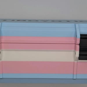 May include: A rectangular storage case with a light blue, pink, and white striped design. The case has two black latches and a sturdy, durable appearance. The color scheme resembles the transgender pride flag.