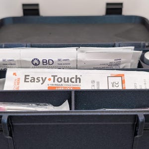 May include: A dark blue medical case, open to reveal organized compartments. Inside are packages labeled "BD" and "Easy-Touch" with syringes. A small, silver-colored vial is also visible. The case is designed for medical supplies.