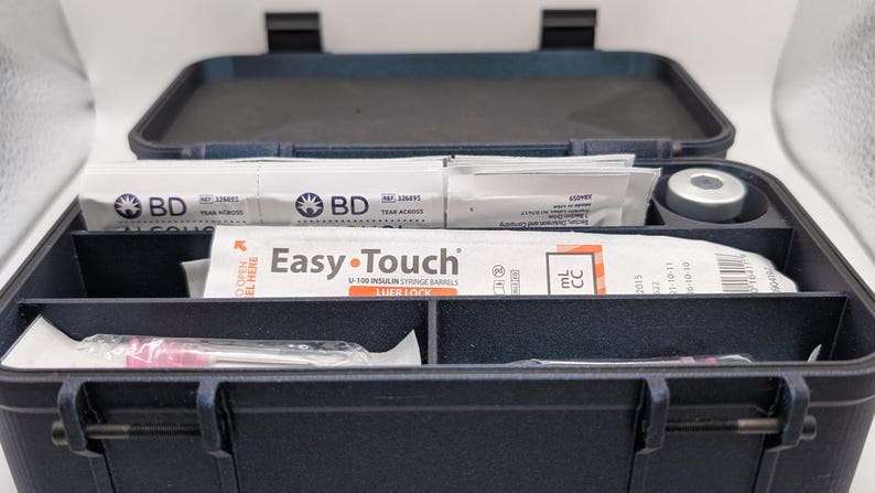 May include: A dark blue medical case, open to reveal organized compartments. Inside are packages labeled "BD" and "Easy-Touch" with syringes. A small, silver-colored vial is also visible. The case is designed for medical supplies.