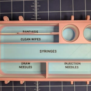 May include: A pink and blue organizer with compartments labeled for medical supplies. The compartments are labeled "BANDAIDS", "CLEAN WIPES", "SYRINGES", "DRAW NEEDLES", and "INJECTION NEEDLES". The organizer has two circular openings.