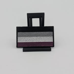May include: A rectangular embroidered patch featuring four horizontal stripes in black, gray, white, and purple. The patch is framed by a thick black border and is displayed on a black plastic stand against a plain, light-colored background.