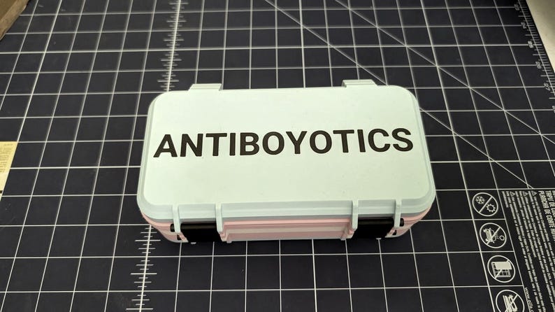 May include: A small, rectangular, light blue case with the word "ANTIBOYOTICS" printed in black on the lid. The case has a pink and black accent and is closed with two black clasps. The case is on a black grid surface.