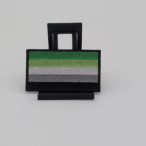 May include: A rectangular embroidered patch featuring horizontal stripes in shades of dark green, light green, white, light gray, and black. The patch is mounted on a black plastic display stand with a square frame and a horizontal base.