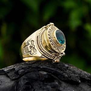 May include: A gold ring with a skull design on the band and a hinged compartment with a green gemstone. The ring is on a dark brown surface.