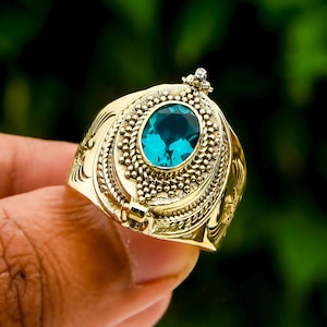 May include: A gold-colored ring with an oval, teal-colored gemstone. The ring has intricate detailing around the gemstone and on the band. The ring is held in a hand against a blurred green background.