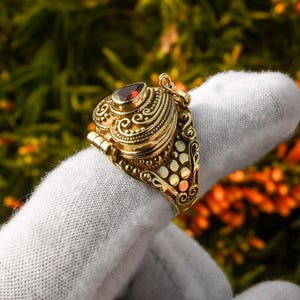 May include: A gold ring with a heart-shaped locket, featuring an intricate design and a red gemstone. The locket opens to reveal a small compartment.