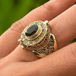 May include: A gold ring with an ornate design and a black oval-shaped stone. The ring has a hinged lid that opens to reveal a small compartment.