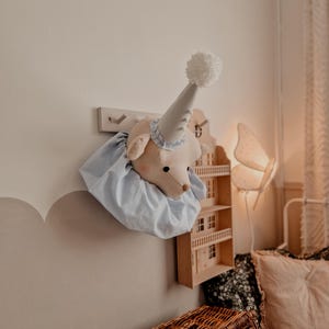 Könnte beinhalten: Ein hellblauer und weißer Stofftierkopf mit einem weißen Bommelhut, der an einer Wand mit einem Holzregal dahinter hängt.
