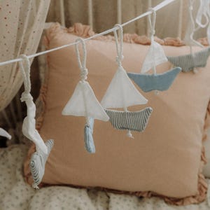 May include: A decorative garland featuring fabric sailboats in shades of blue and white. The boats hang against a peach-coloured pillow. The sails are white, and the hulls are striped or solid blue. A floral bedspread and sheer curtain are in the background.
