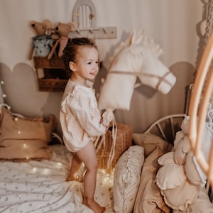 Puede incluir: Una ni&ntilde;a juega con un caballo de peluche blanco en una habitaci&oacute;n con una cama blanca, ropa de cama floral y varias almohadas.