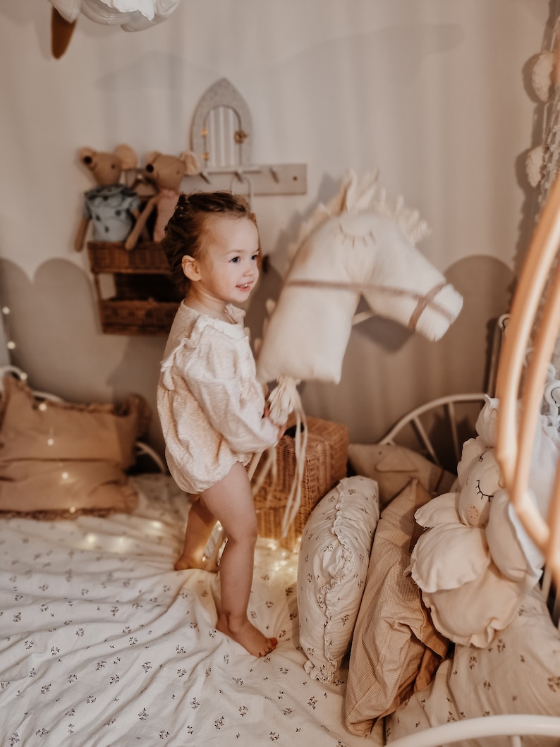 Puede incluir: Una ni&ntilde;a juega con un caballo de peluche blanco en una habitaci&oacute;n con una cama blanca, ropa de cama floral y varias almohadas.