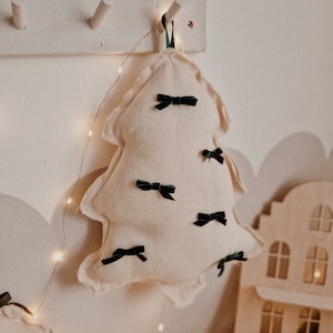 May include: A white fabric Christmas tree ornament with green velvet bows hanging from a hook on a white wall. The ornament is decorated with a string of white fairy lights.
