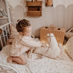 Puede incluir: Una ni&ntilde;a peque&ntilde;a con un vestido rosa y blanco se sienta en una cama con una s&aacute;bana de estampado floral blanco. Est&aacute; jugando con un animal de peluche blanco con forma de cisne. La cama est&aacute; decorada con luces de hadas y una maleta de mimbre.