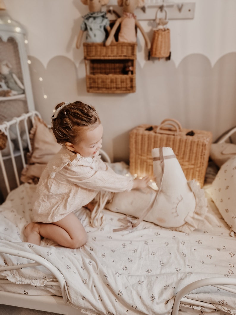 Puede incluir: Una ni&ntilde;a peque&ntilde;a con un vestido rosa y blanco se sienta en una cama con una s&aacute;bana de estampado floral blanco. Est&aacute; jugando con un animal de peluche blanco con forma de cisne. La cama est&aacute; decorada con luces de hadas y una maleta de mimbre.