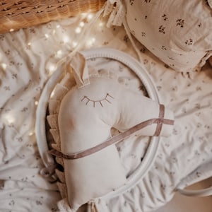 Puede incluir: Una cabeza de caballo de peluche blanca con una correa de tela marr&oacute;n y pesta&ntilde;as bordadas. La cabeza de caballo est&aacute; sentada sobre una tela blanca con un patr&oacute;n floral.