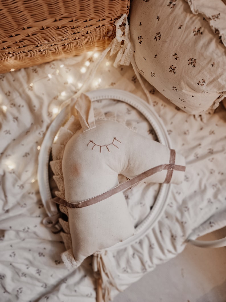 Puede incluir: Una cabeza de caballo de peluche blanca con una correa de tela marr&oacute;n y pesta&ntilde;as bordadas. La cabeza de caballo est&aacute; sentada sobre una tela blanca con un patr&oacute;n floral.