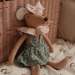 Puede incluir: Una muñeca de ratón de peluche con un lazo rosa, un cuello con volantes y un vestido floral verde. La muñeca está sentada sobre una manta estampada. Una cesta de mimbre está en el fondo.