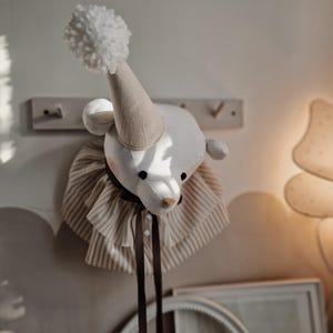 Puede incluir: Una cabeza de animal de peluche blanca con un sombrero de pompón blanco y un cuello a rayas blanco y marrón. La cabeza está montada en una pared con una cinta marrón que cuelga.
