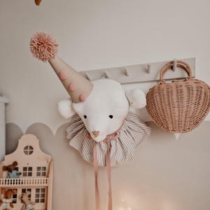 Può includere: Una testa di animale di peluche bianca con un cappello a pois rosa e bianco e un fiocco rosa. La testa è montata su una parete con un cestino di vimini a forma di cuore marrone appeso accanto.