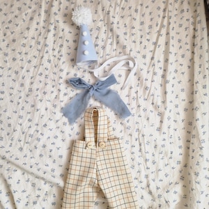 May include: A baby outfit set, including a light blue party hat with a white pom-pom and three white buttons, a matching blue bow tie, and plaid overalls with wooden buttons. The items are arranged on a floral patterned fabric.