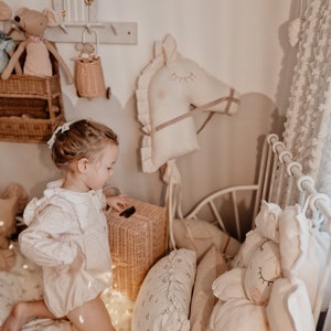 Puede incluir: Una ni&ntilde;a peque&ntilde;a con un vestido blanco juega en una habitaci&oacute;n con un caballo de balanc&iacute;n blanco, una cesta de mimbre y un animal de peluche. La habitaci&oacute;n est&aacute; decorada en un esquema de color neutro con paredes y ropa de cama blancas.