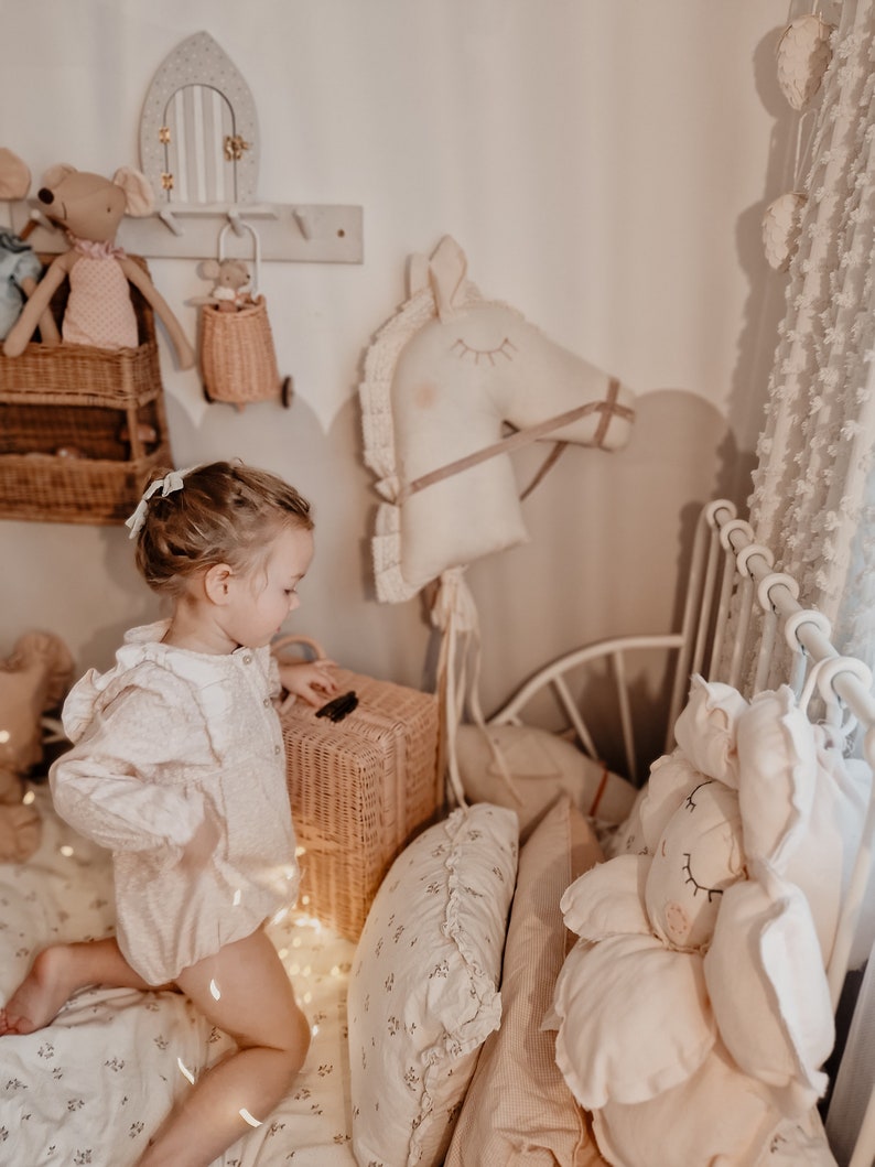 Puede incluir: Una ni&ntilde;a peque&ntilde;a con un vestido blanco juega en una habitaci&oacute;n con un caballo de balanc&iacute;n blanco, una cesta de mimbre y un animal de peluche. La habitaci&oacute;n est&aacute; decorada en un esquema de color neutro con paredes y ropa de cama blancas.