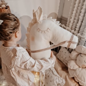 Puede incluir: Una ni&ntilde;a peque&ntilde;a juega con un juguete de caballo de balanc&iacute;n de peluche blanco con una silla de montar y riendas marrones. El caballo tiene ojos bordados y un borde de encaje. La ni&ntilde;a lleva un vestido blanco con flores.