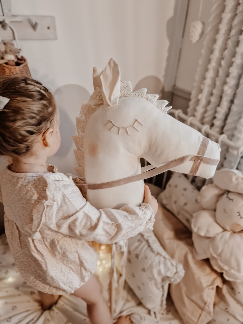 Puede incluir: Una ni&ntilde;a peque&ntilde;a juega con un juguete de caballo de balanc&iacute;n de peluche blanco con una silla de montar y riendas marrones. El caballo tiene ojos bordados y un borde de encaje. La ni&ntilde;a lleva un vestido blanco con flores.