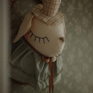 May include: A plush bunny head with a plaid hat and a striped collar. The bunny has closed eyes with long lashes and a brown nose. A brown ribbon is tied around the neck. The background is a floral wallpaper.